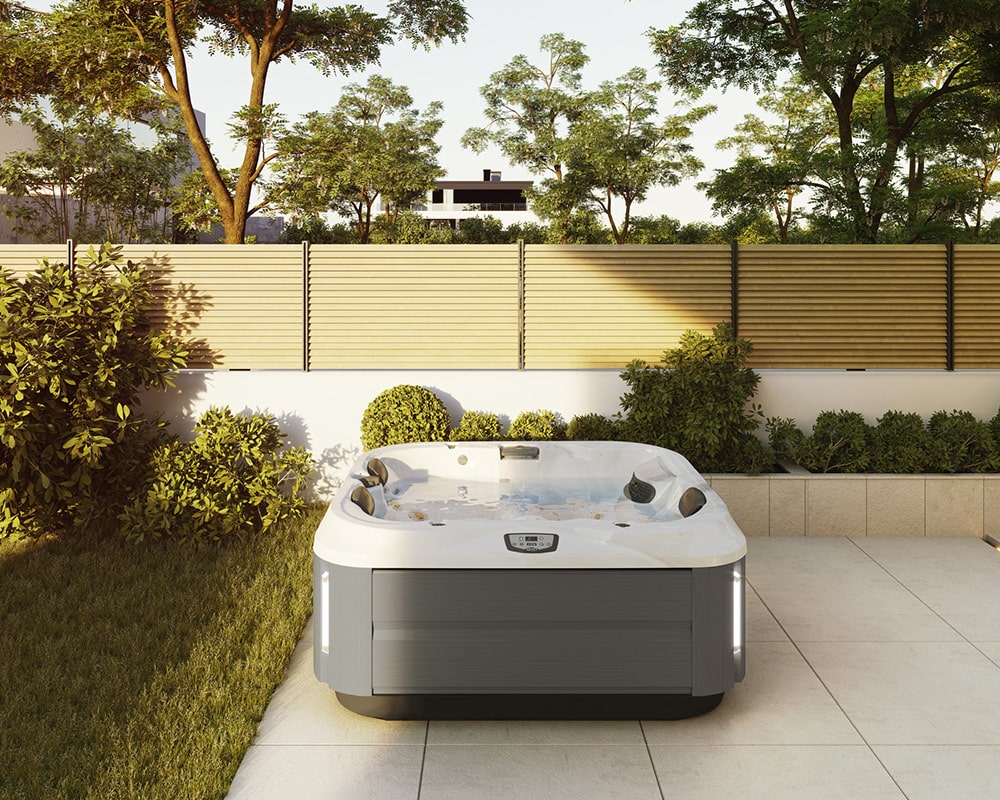 A modern JHT-J315 plug & play hot tub filled with water is placed outdoors, surrounded by a well-maintained garden and a fence